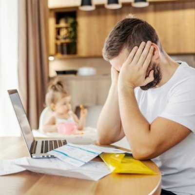 a man sits at a table with his face in his hands, in front of him are papers and a laptop, behind him is a small child in a high chair 